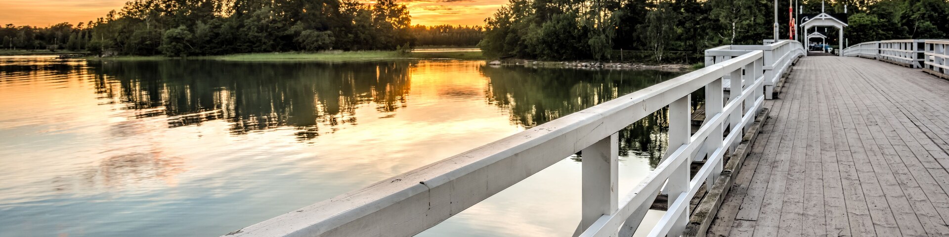 A wooden pier extending over calm water, with trees and a sunset in the background.
