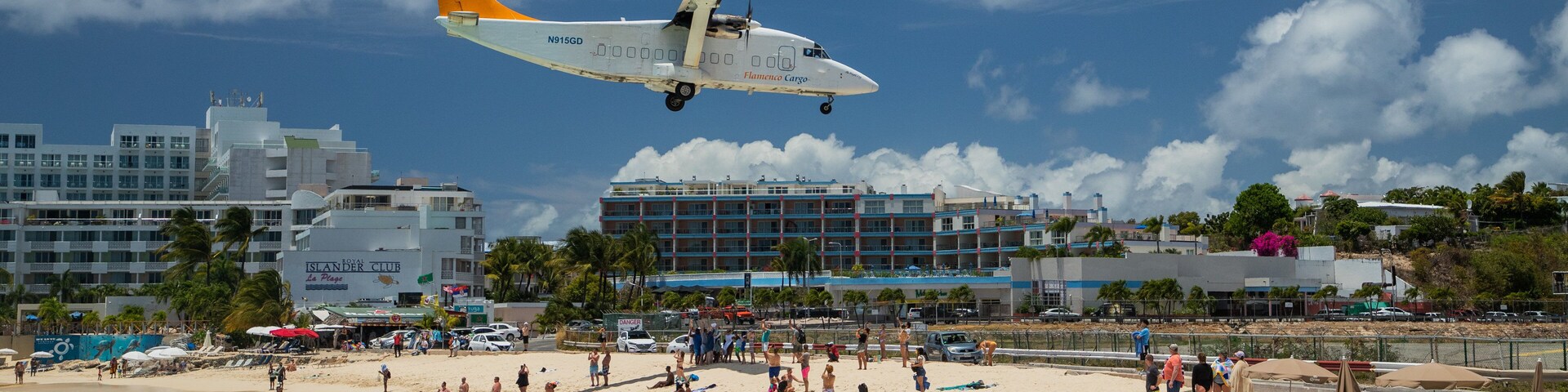 Ein Strand mit Menschen, ein niedrig fliegendes Flugzeug und Gebäude im Hintergrund.