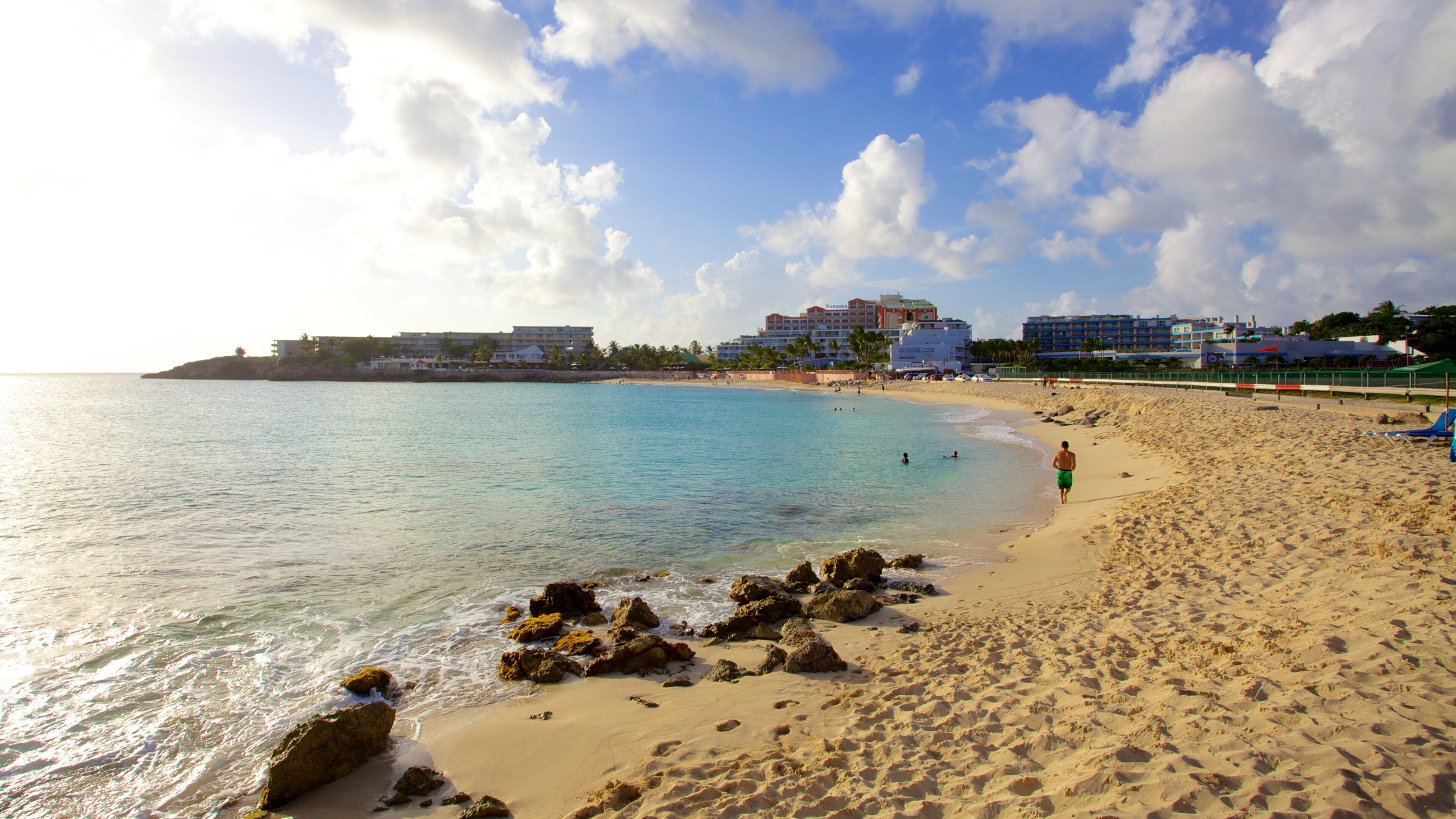 A beach with clear turquoise water, sandy shore, and buildings in the background.