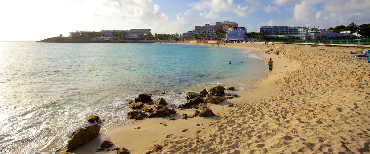 A beach with clear turquoise water, sandy shore, and buildings in the background.
