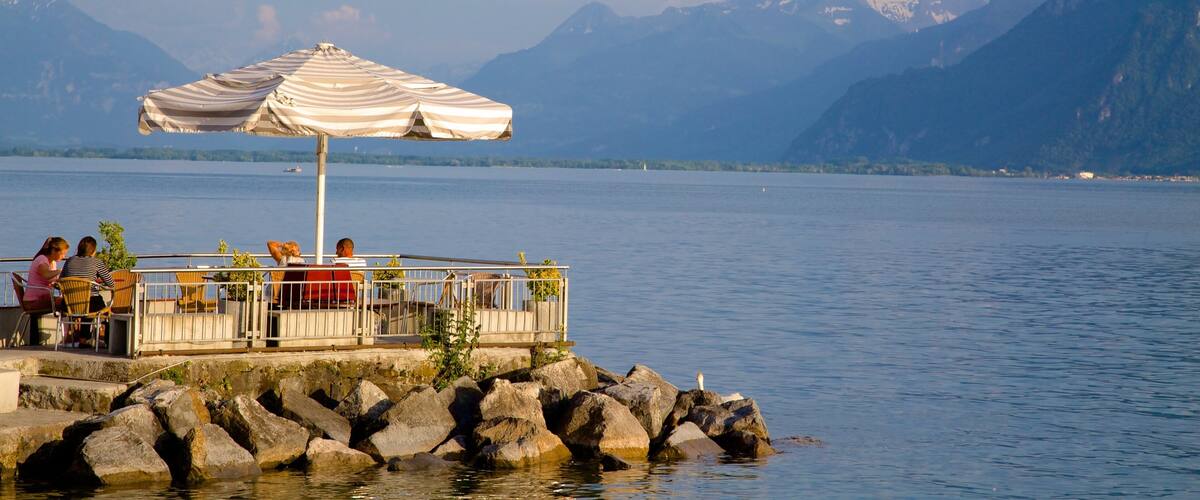 A lakeside terrace with a white umbrella, people sitting, and mountains in the background.