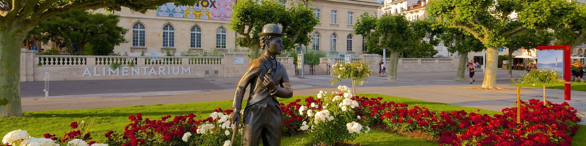 A statue in a garden with red and white flowers, surrounded by a paved area and buildings in the background.