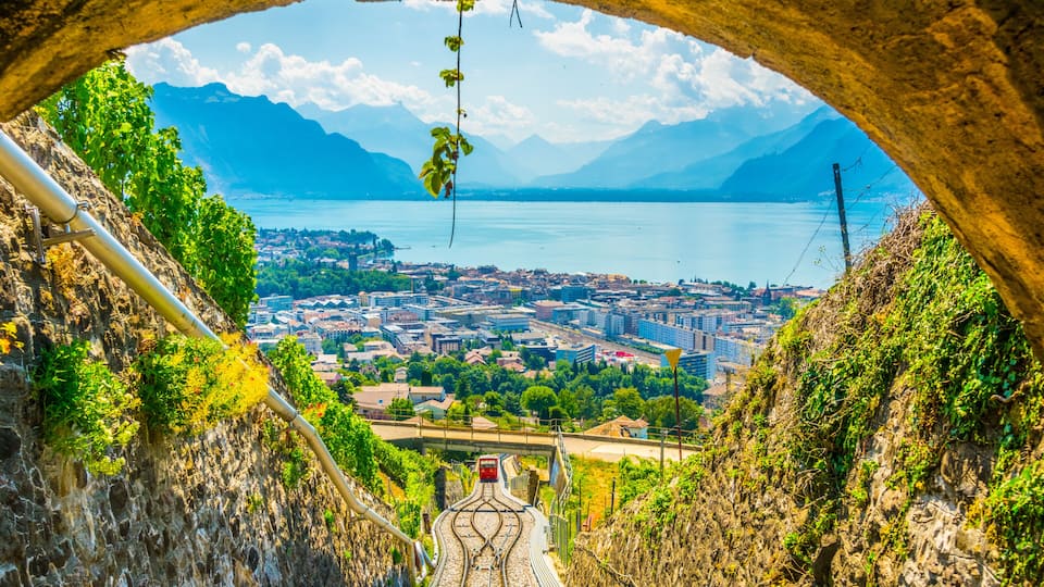 A mountain railway track leading to a town with a lake and mountains in the background.