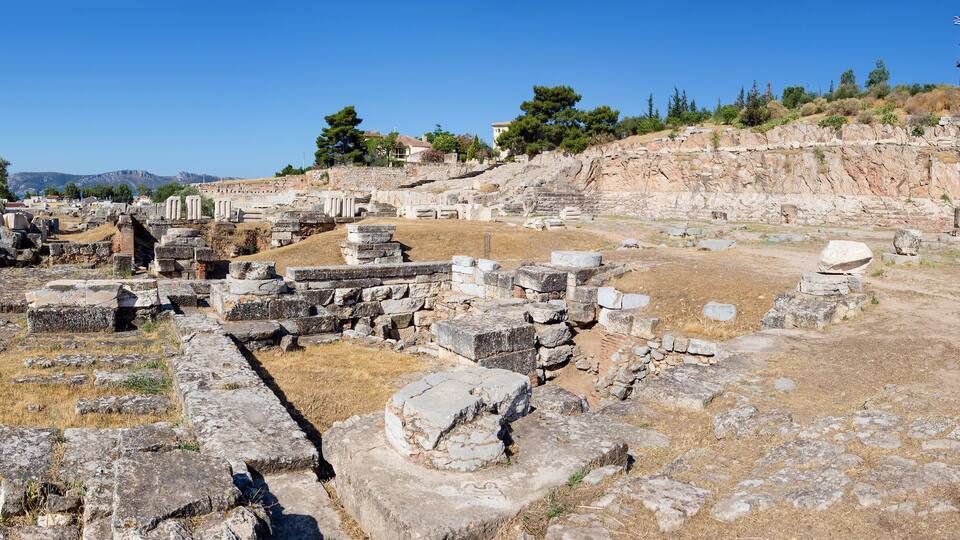 Ancient ruins with stone blocks and columns, a watchtower, and a hillside with trees.