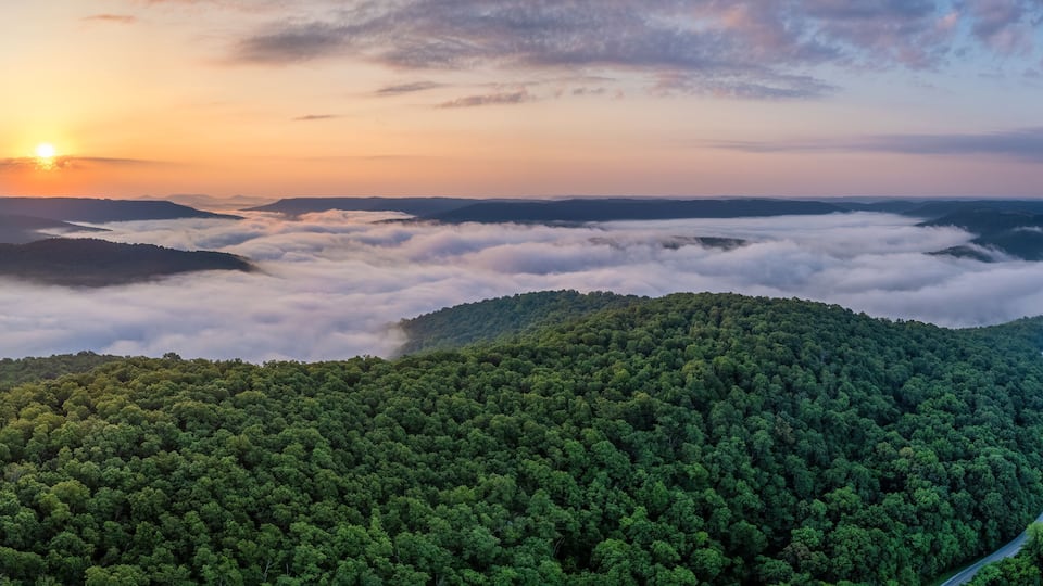 A forested mountain range with a winding road and a sunrise.