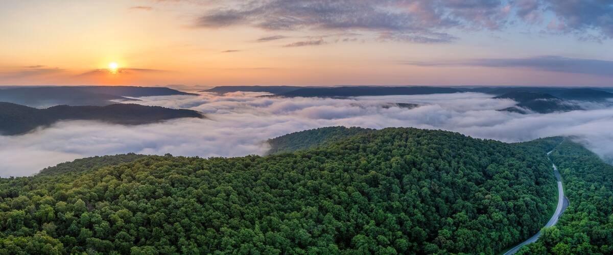 A forested mountain range with a winding road and a sunrise.