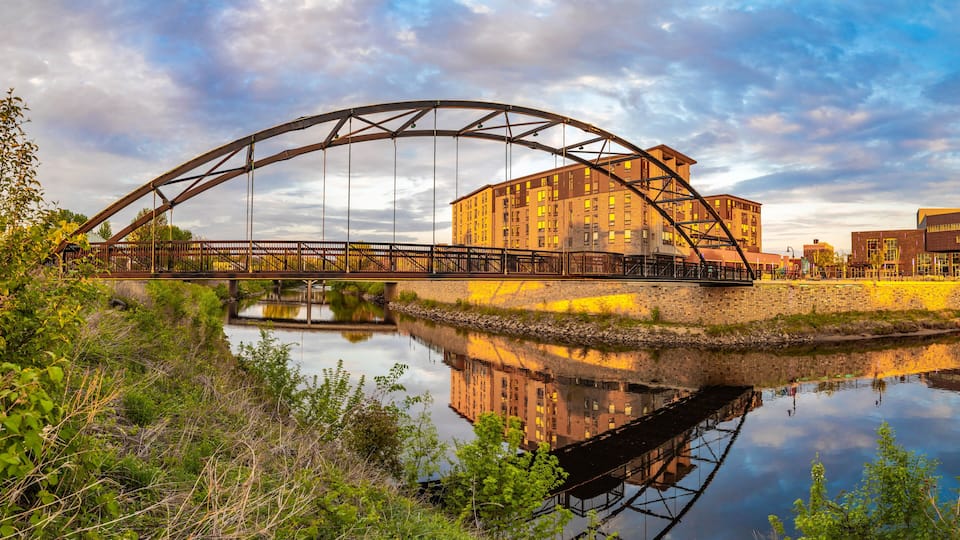 A steel bridge over a calm river, reflecting a building and the sky.