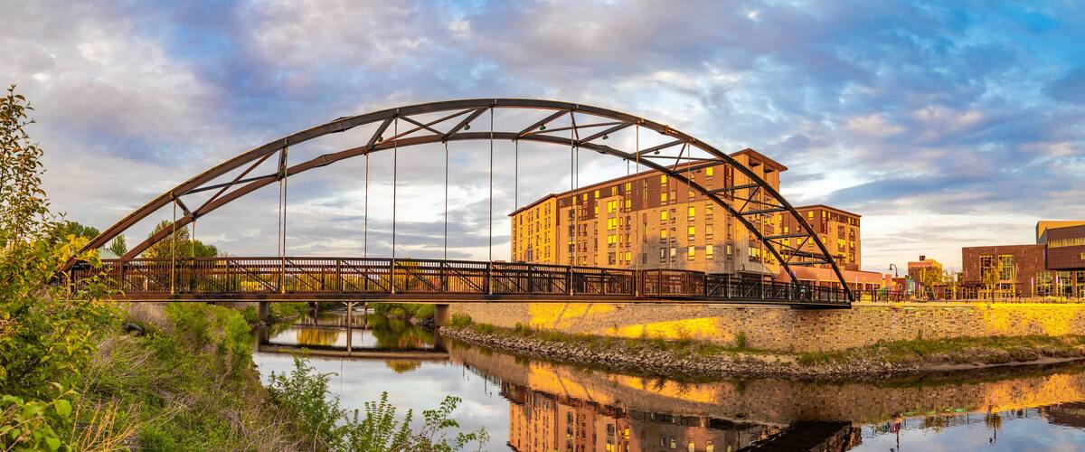 A steel bridge over a calm river, reflecting a building and the sky.