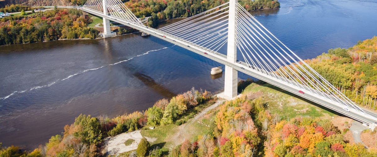 Un puente colgante sobre un río con follaje otoñal y un pueblo lejano.