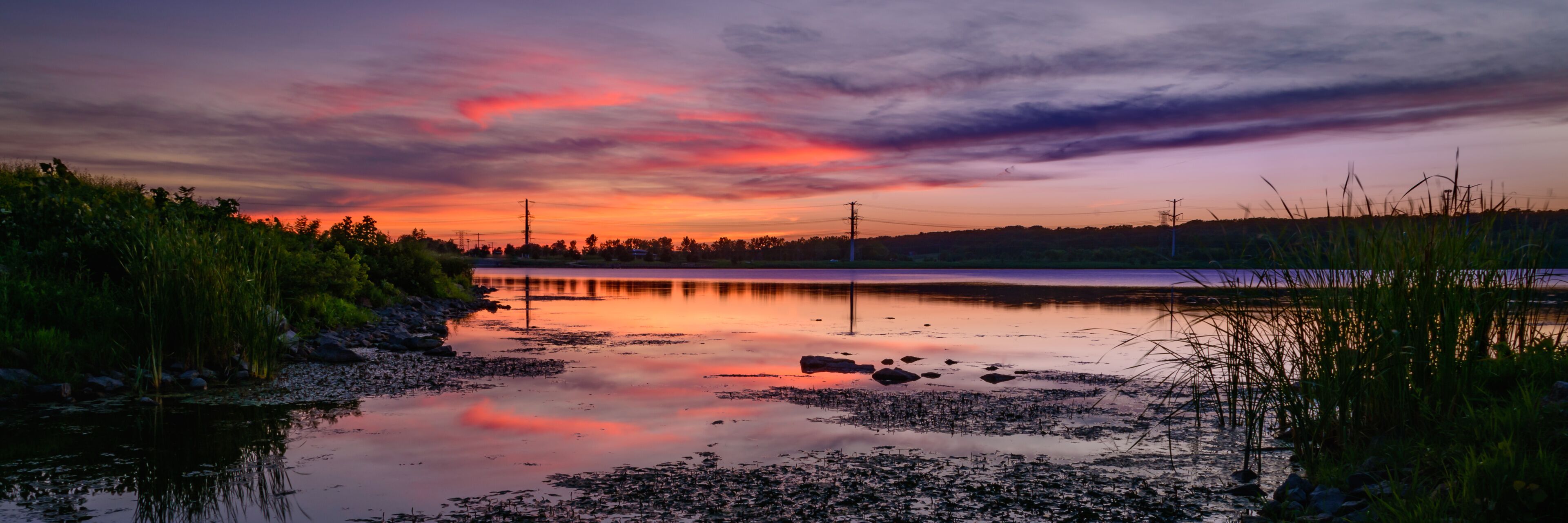 A sunset over a calm lake with reflections, surrounded by vegetation and power lines.