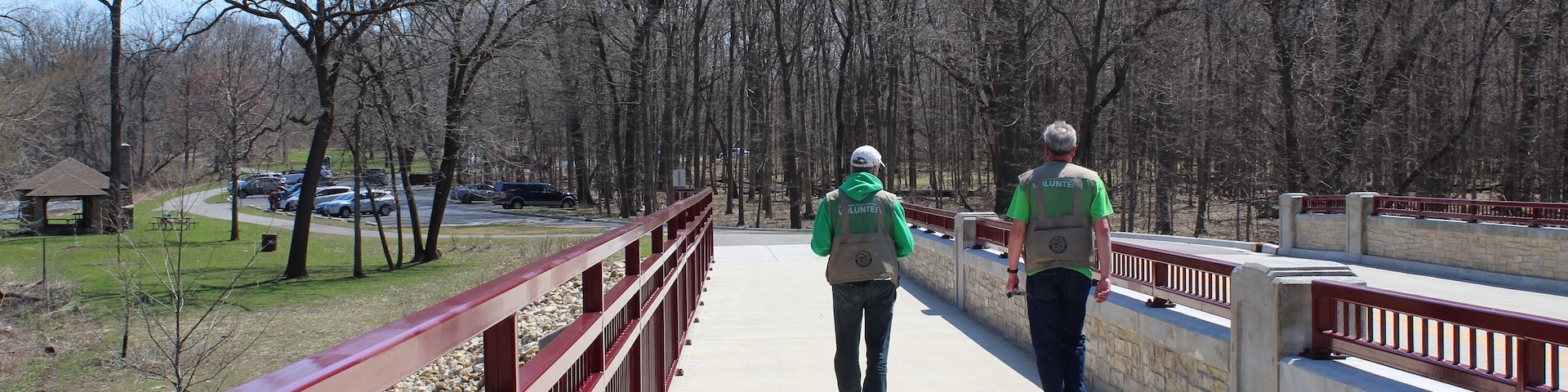 Duas pessoas caminhando sobre uma ponte com guarda-corpo vermelho, cercadas por árvores e um céu limpo.