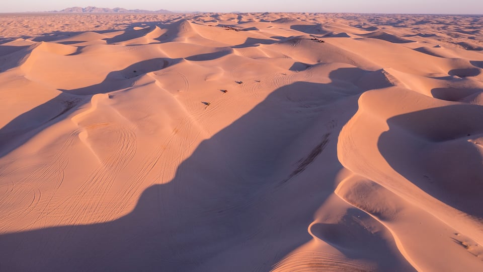 A vast desert landscape with undulating sand dunes and distant mountain ranges.