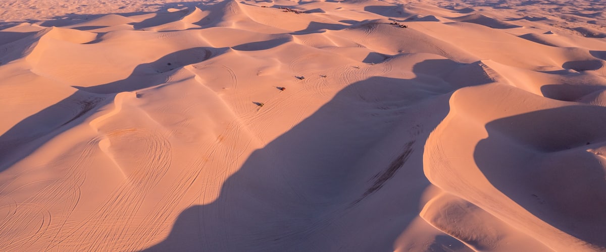 A vast desert landscape with undulating sand dunes and distant mountain ranges.