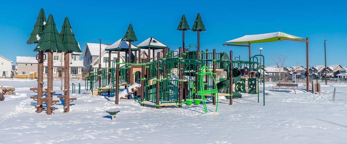 A snow-covered playground with green climbing structures and a yellow umbrella.