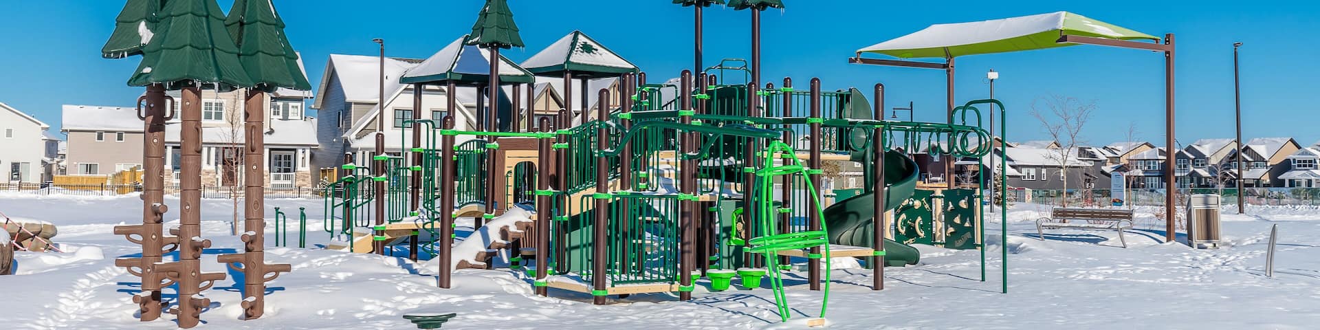 A snow-covered playground with green climbing structures and a yellow umbrella.