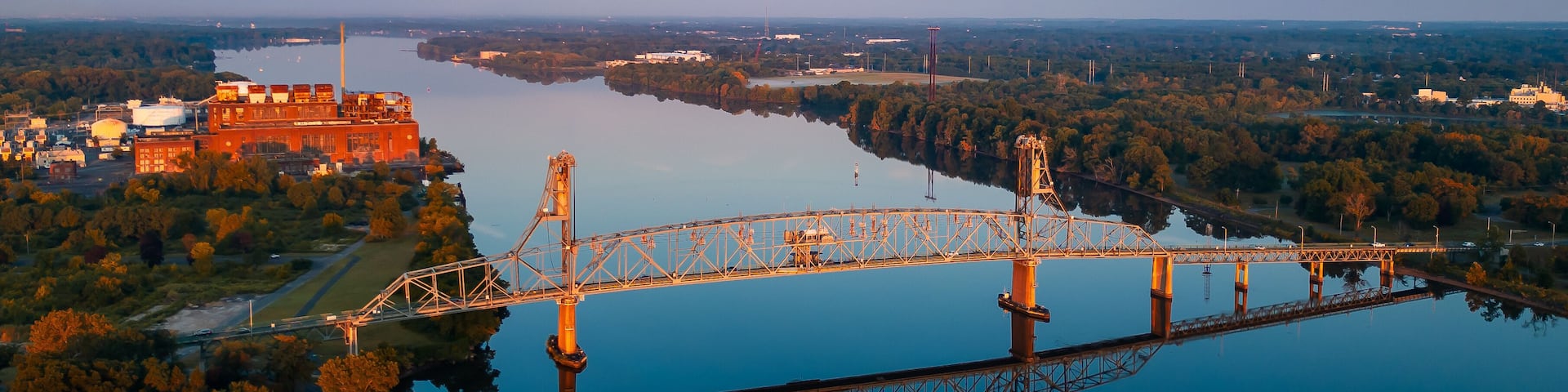 A bridge over a calm river, surrounded by trees and industrial buildings.