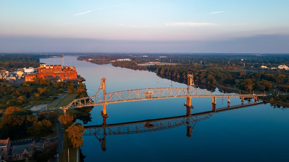 A bridge over a calm river, surrounded by trees and industrial buildings.