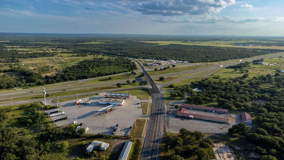 An aerial view of a highway interchange with multiple lanes, surrounded by green fields and sparse buildings.