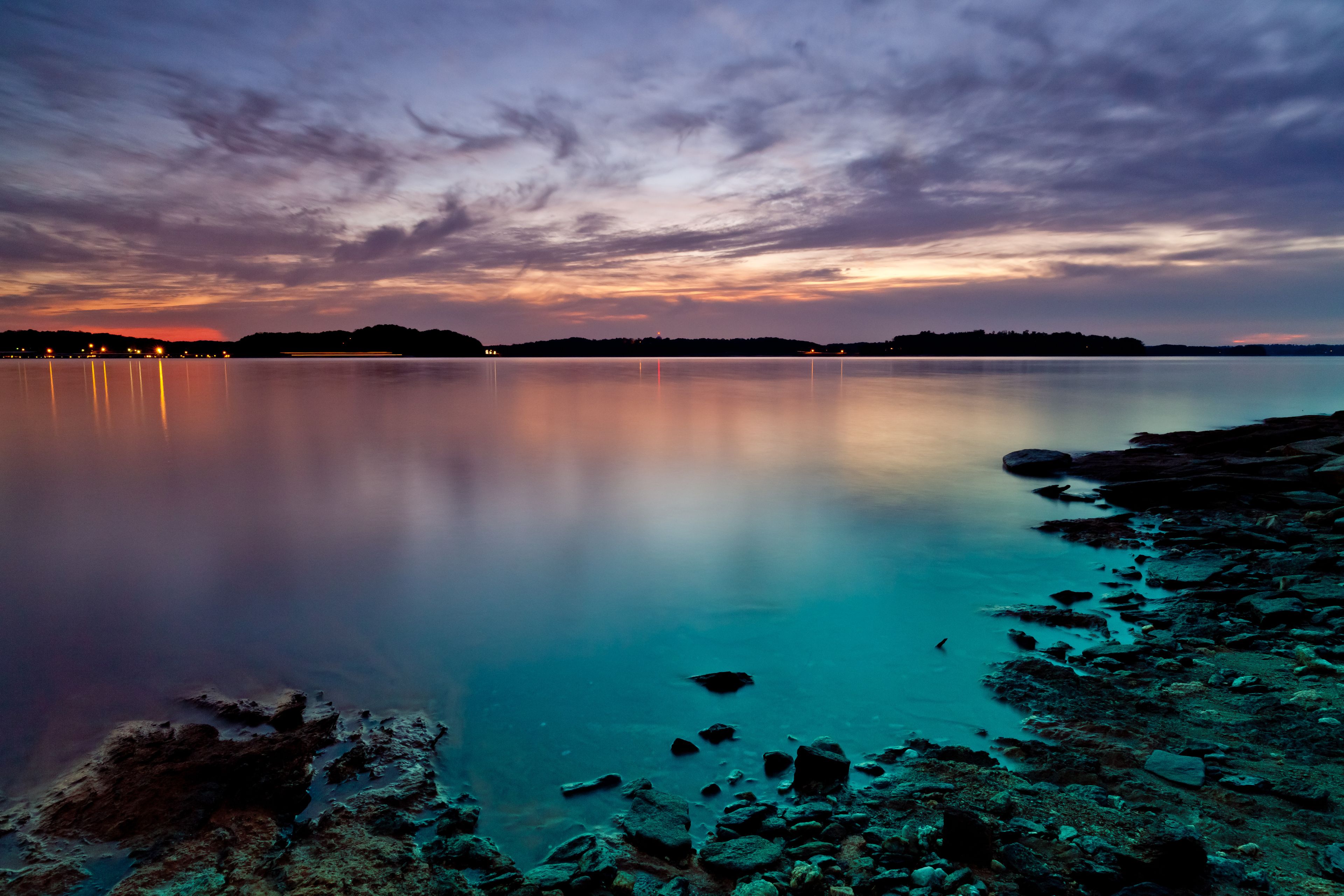 A calm lake at dusk with a rocky shoreline and a distant city skyline.