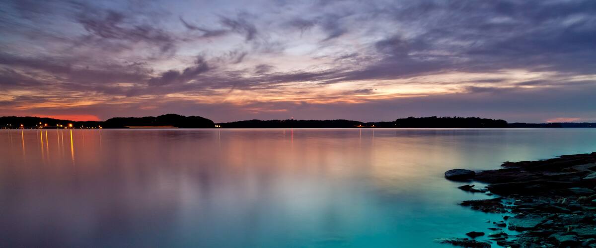 A calm lake at dusk with a rocky shoreline and a distant city skyline.
