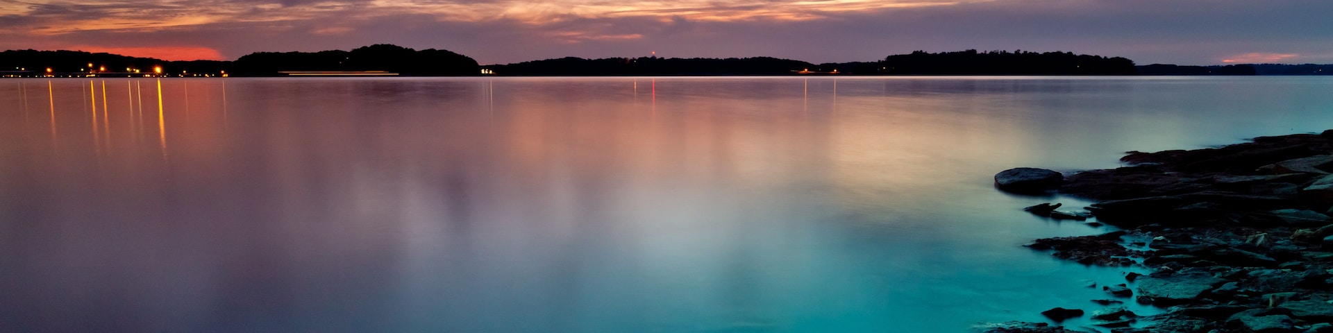 Ein ruhiger See im Abenddämmerung mit steiniger Uferlinie und einer entfernten Stadtansicht.