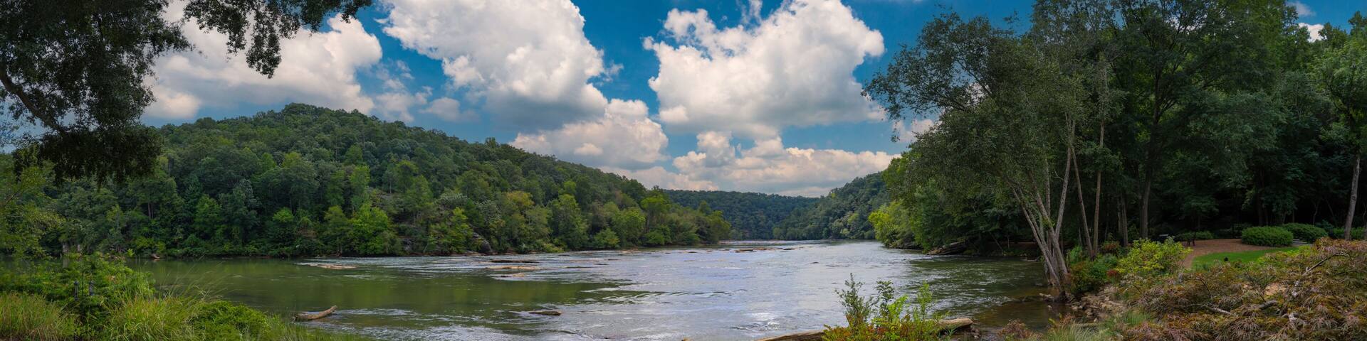 a panoramic of a gorgeous summer landscape on the Chattahoochee River with flowing water surrounded by lush green trees, grass and plants and blue sky with clouds at Walton On The Chattahoochee