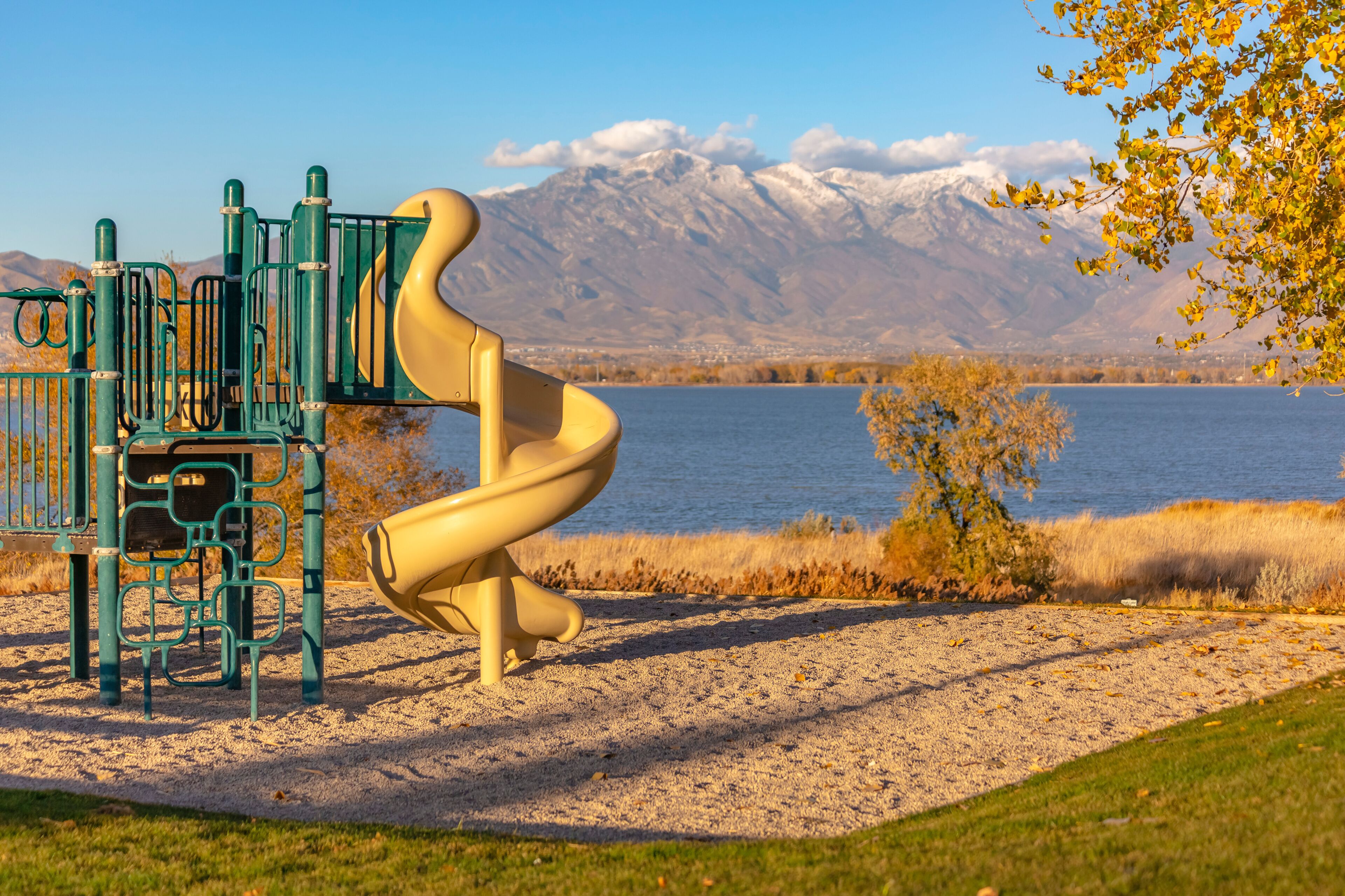 Un parque infantil con tobogán y estructuras para escalar, una zona de arena y un lago con montañas al fondo.