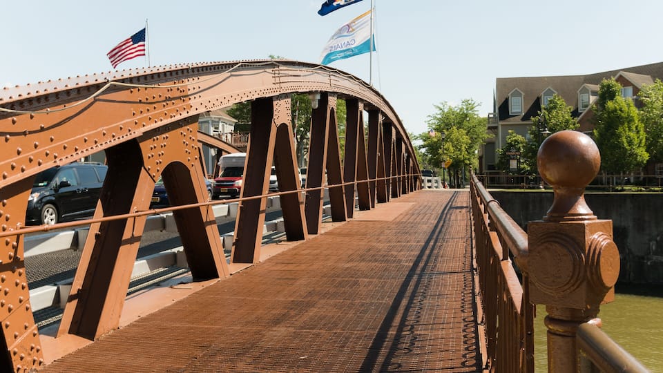 A brown steel bridge with American and Canadian flags, a metal railing, and a building in the background.