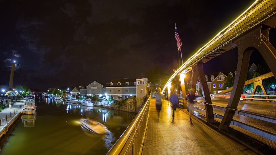 A nighttime view of a bridge over a calm river with boats docked on the left, a lit walkway, and residential buildings in the background.