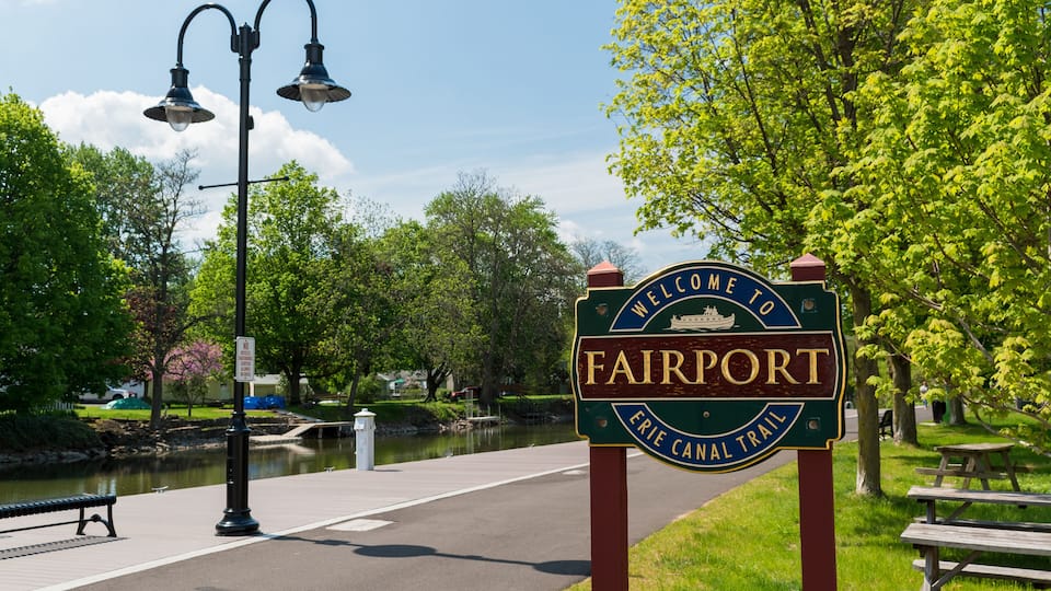 A welcome sign for Fairport on Erie Canal Trail, with a bench and lamp post nearby.