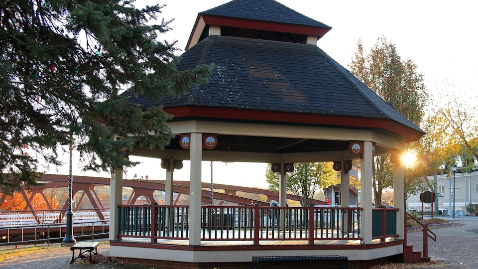 A gazebo with a red roof and white walls, surrounded by trees and a bridge in the background.