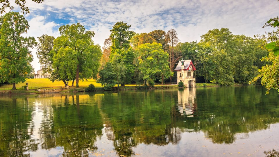 A serene lake with a red-roofed house surrounded by lush greenery and trees.