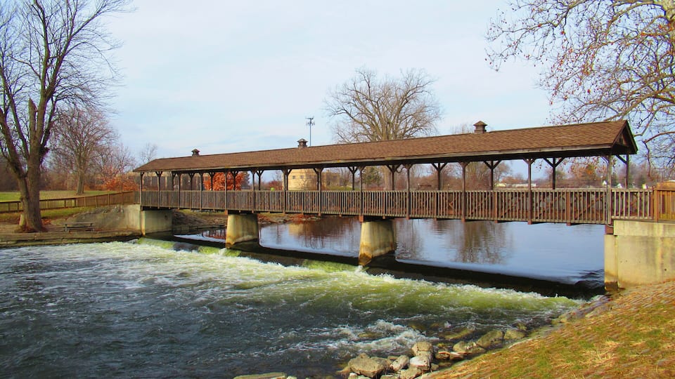 A wooden covered bridge over a flowing river with rocks in the foreground and trees in the background.