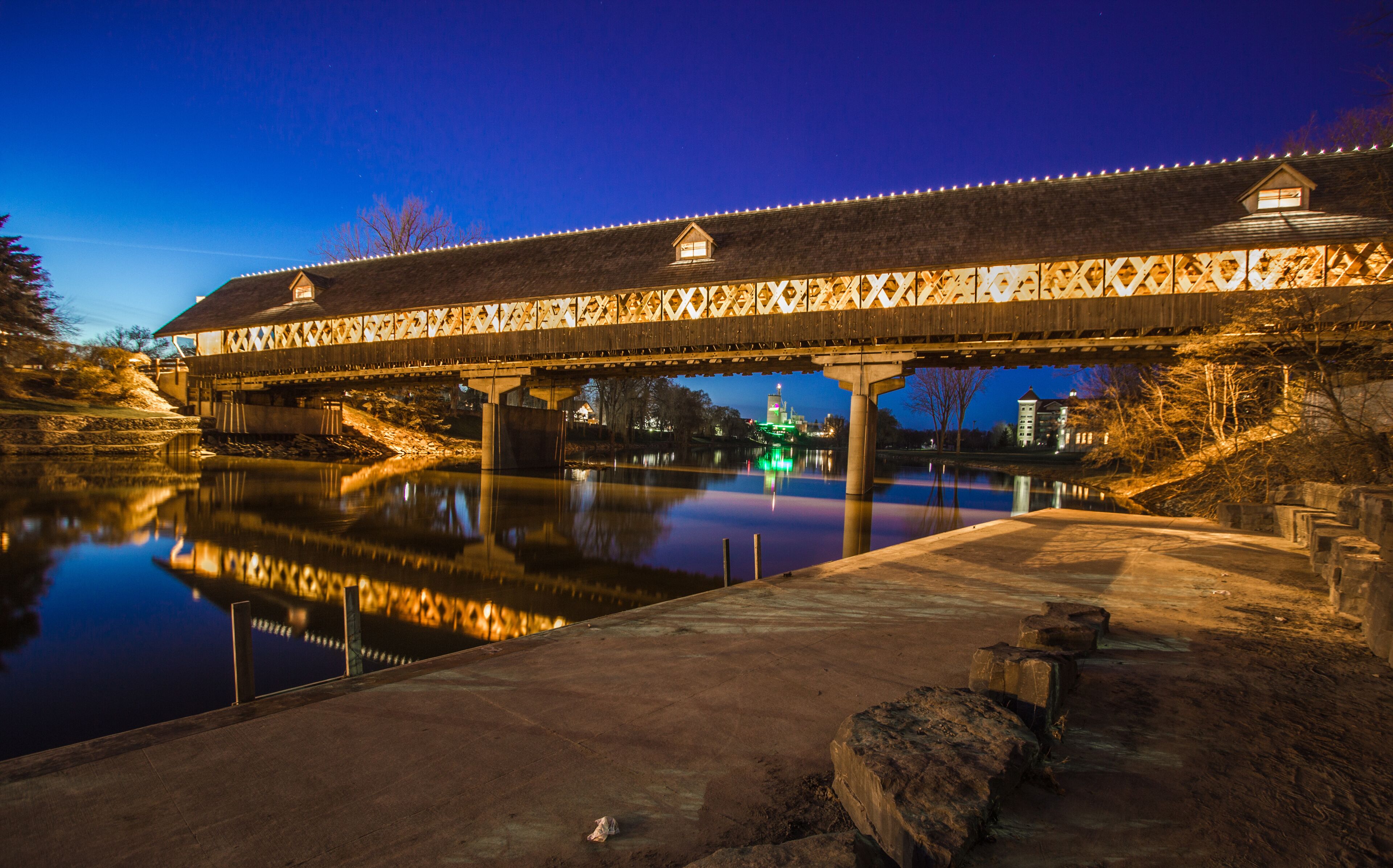 A covered bridge at dusk with lights, a calm river below, and a building with a green light in the background.