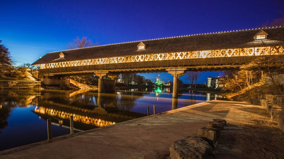 A covered bridge at dusk with lights, a calm river below, and a building with a green light in the background.