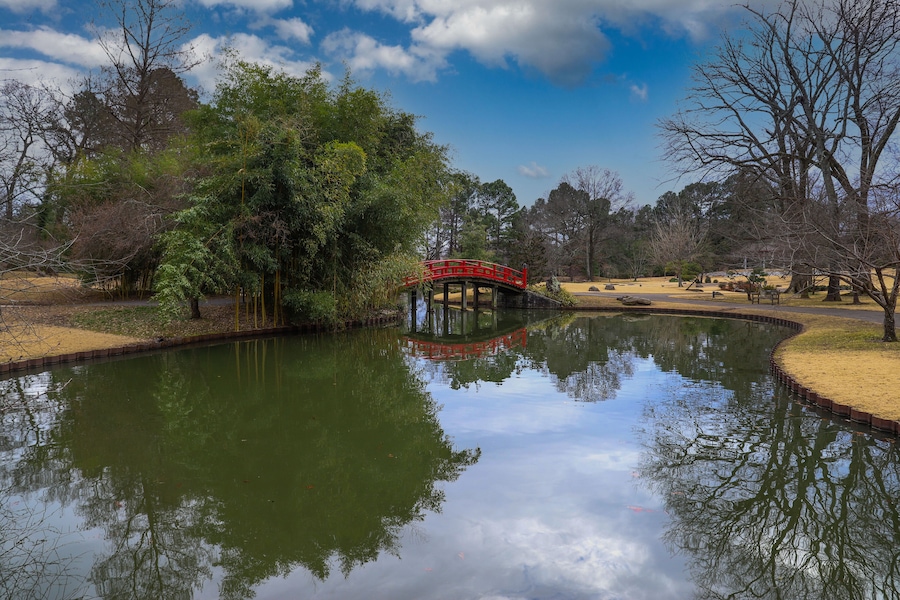 a shot of an arched red and black bridge over a lake surrounded lush green trees reflecting off the water with blue sky and powerful clouds at Memphis Botanic Garden in Memphis Tennessee USA