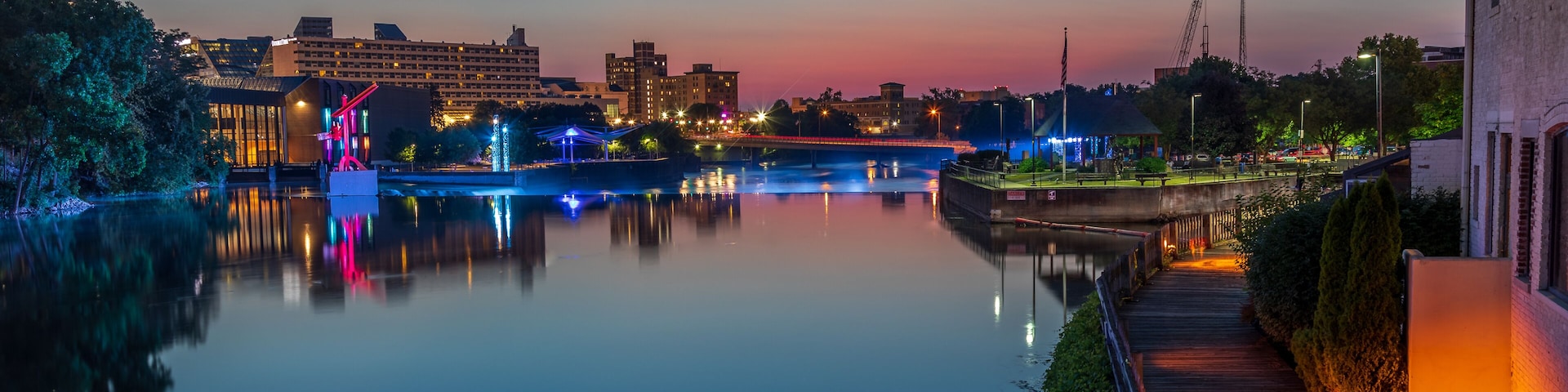 A calm river at dusk with illuminated structures and a bridge.