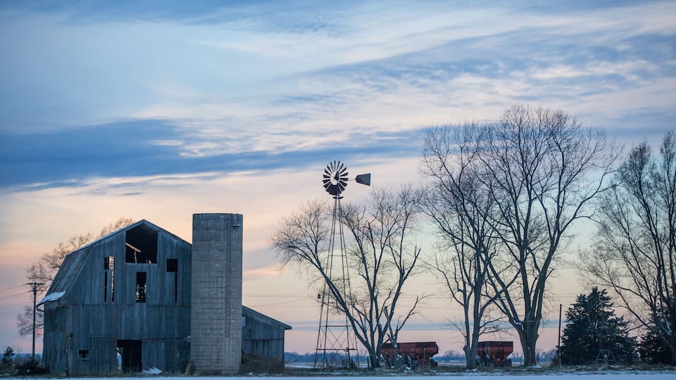 A barn, a windmill, and leafless trees in a snowy field.