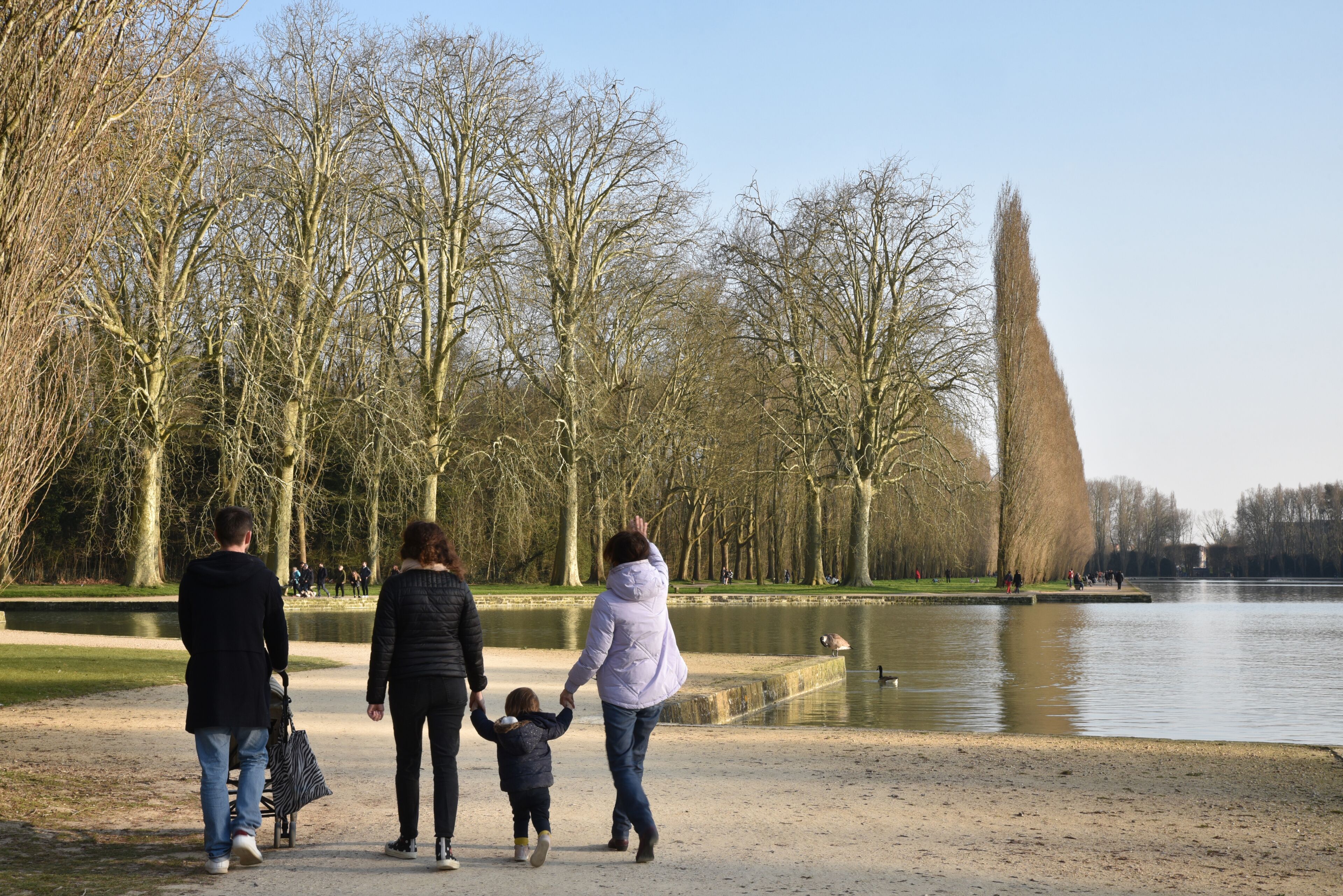 Una familia caminando por un sendero junto al lago en un parque con árboles sin hojas y una torre a lo lejos.