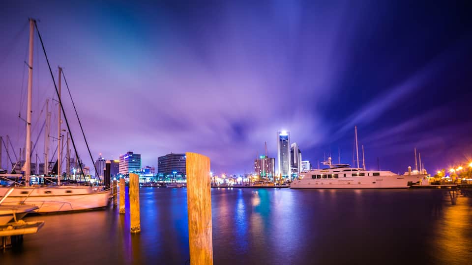 A marina at night with boats docked, a large yacht, and illuminated buildings in the background.