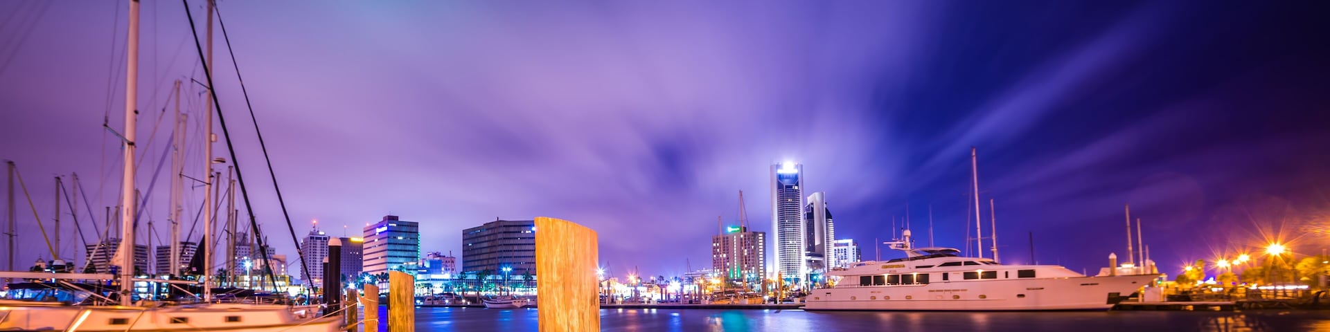A marina at night with boats docked, a large yacht, and illuminated buildings in the background.