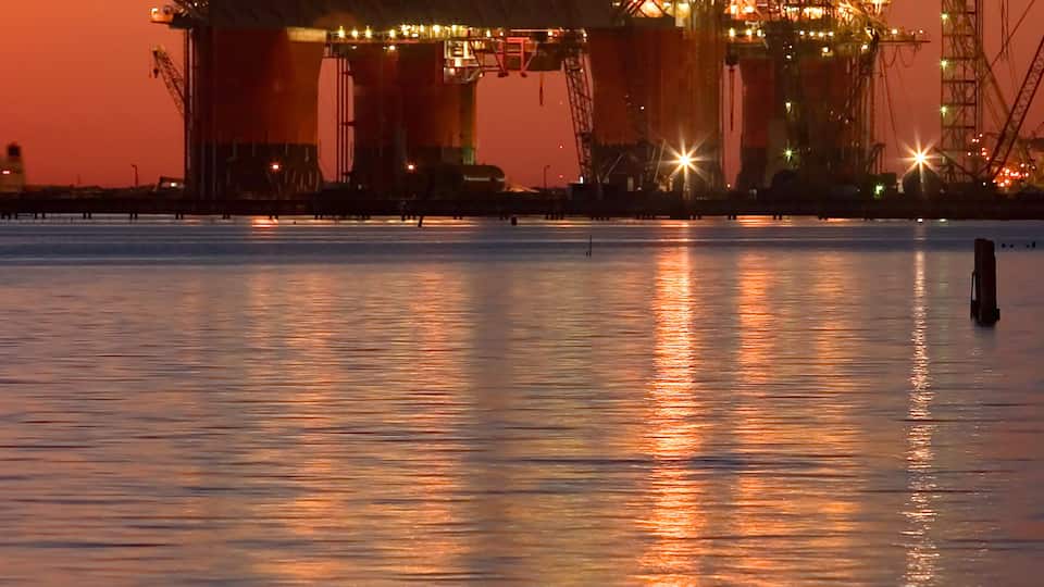 An offshore oil platform at sunset with cranes and lights reflecting on the water.