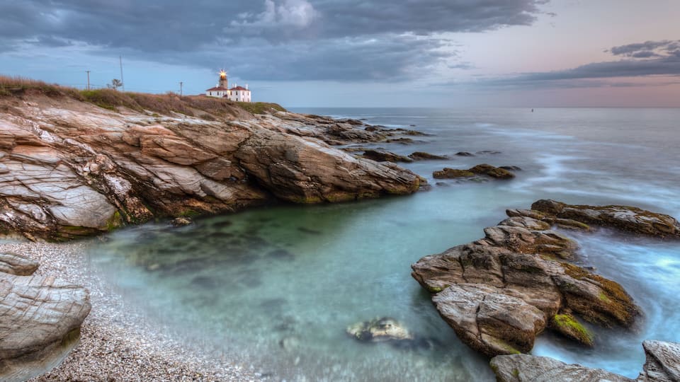 A coastal landscape with a lighthouse, rocky shoreline, and clear water.