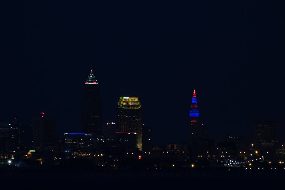 A city skyline at night with illuminated buildings, including a prominent tower with a red and blue top.