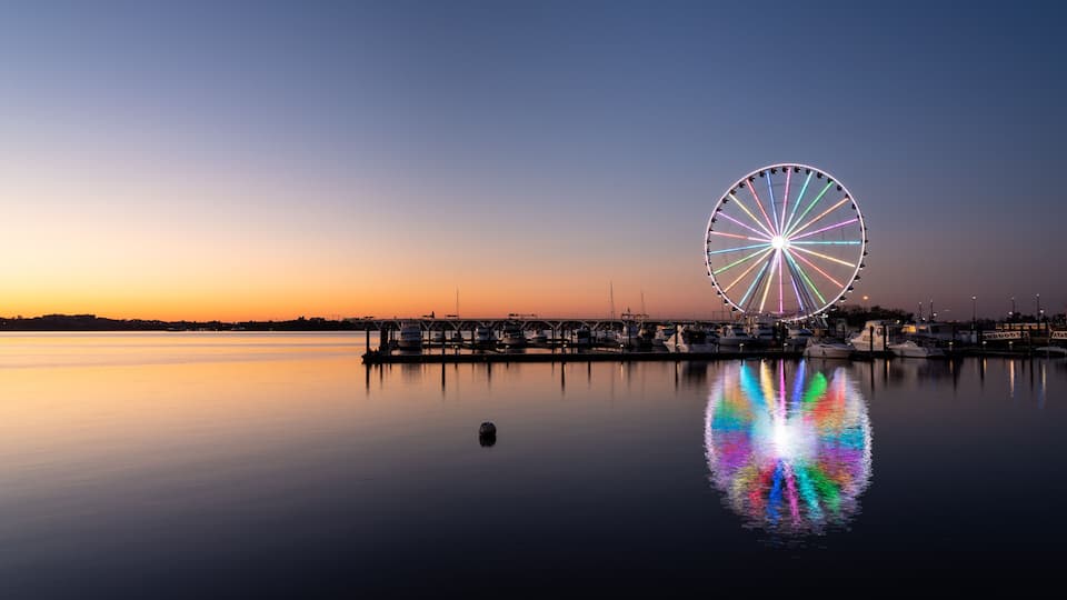 A calm waterfront at sunset with a large illuminated Ferris wheel reflecting on the water.