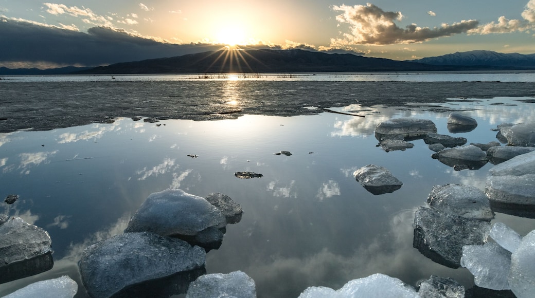 A frozen lake with ice chunks in the foreground and the sun setting behind mountains.