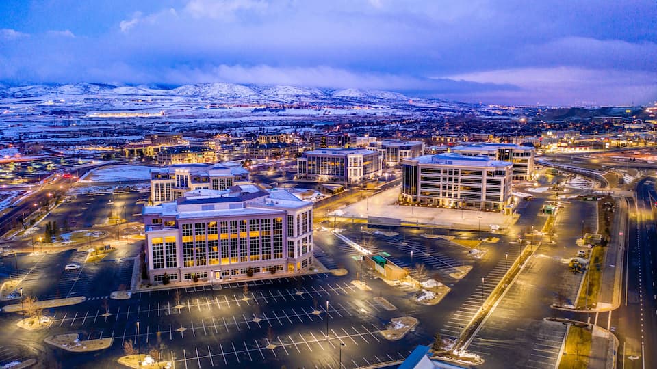 An aerial view of a city with modern buildings, a snow-covered mountain range, and a large parking area.