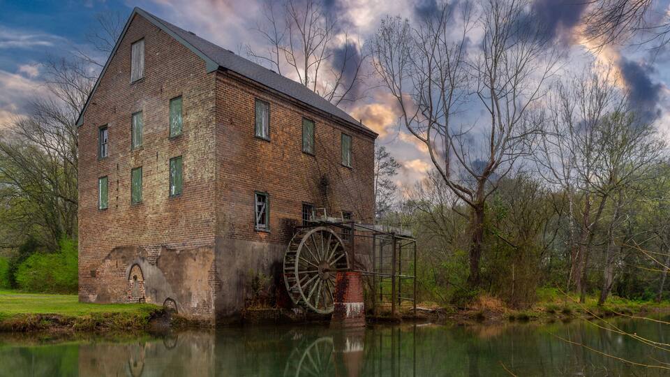 A historic brick mill with a large water wheel, surrounded by trees and a calm river.