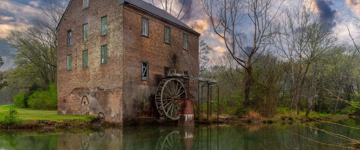 A historic brick mill with a large water wheel, surrounded by trees and a calm river.