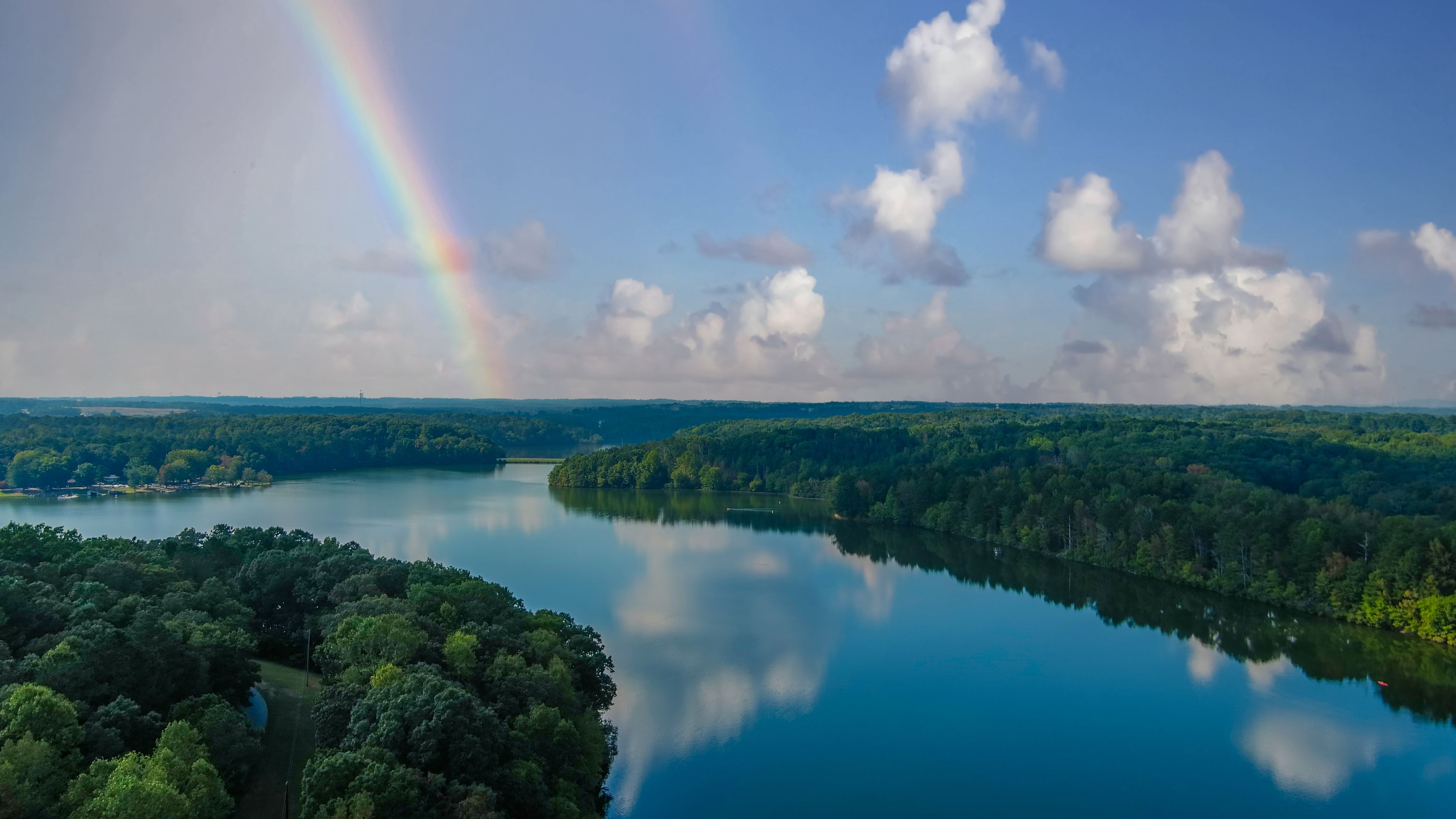 stunning aerial shot of the still waters of the lake with blue sky and clouds reflecting off the water and of lush green trees and a rainbow at Sweetwater Creek State Park in Lithia Springs Georgia